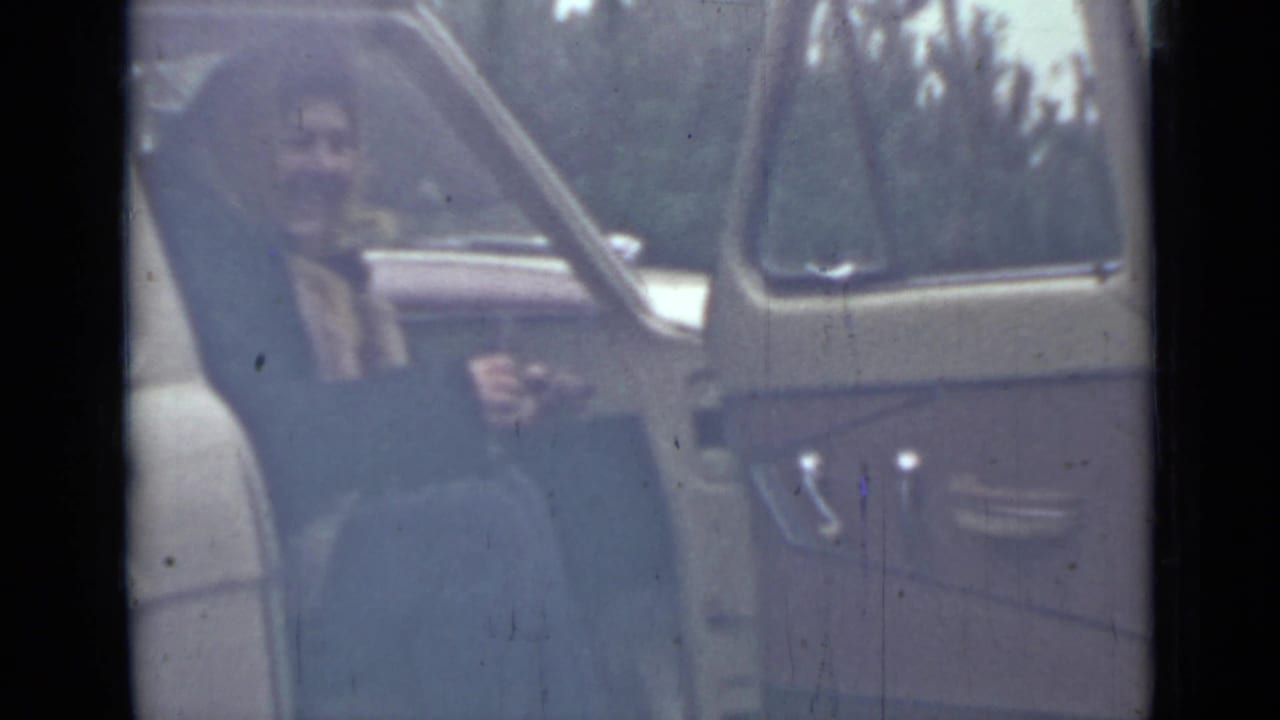 Woman seated beside an open yellow car door in soft 8mm footage.