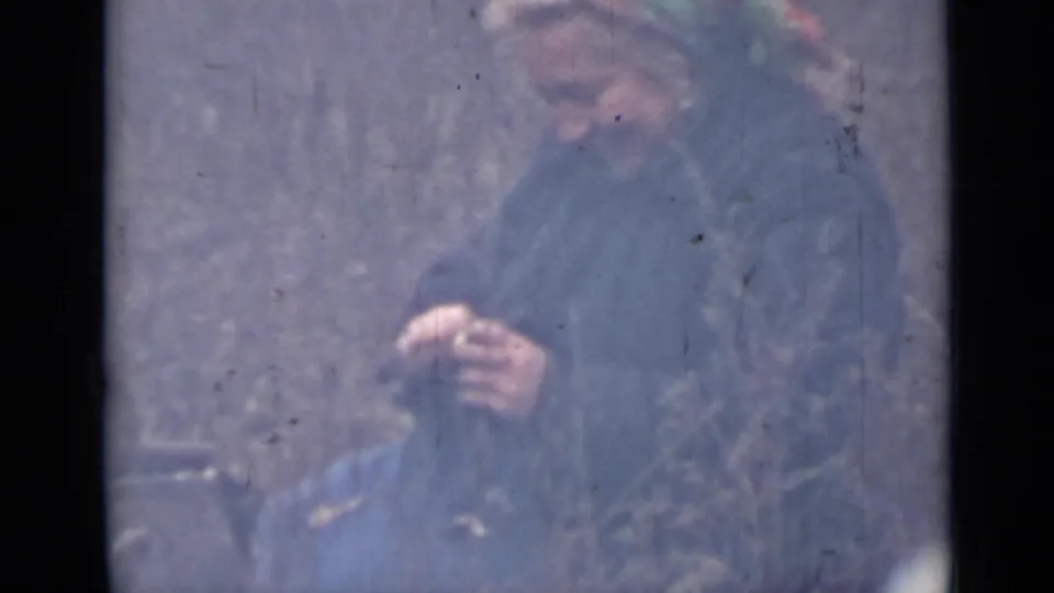 In a faded 8mm frame, the woman remains focused on a small peeling task outdoors, with her headscarf and coat visible.