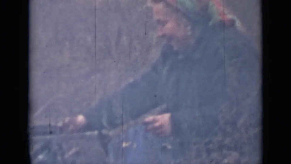 The woman in a headscarf shifts position while working with food and a knife, seen in grainy color 8mm footage.