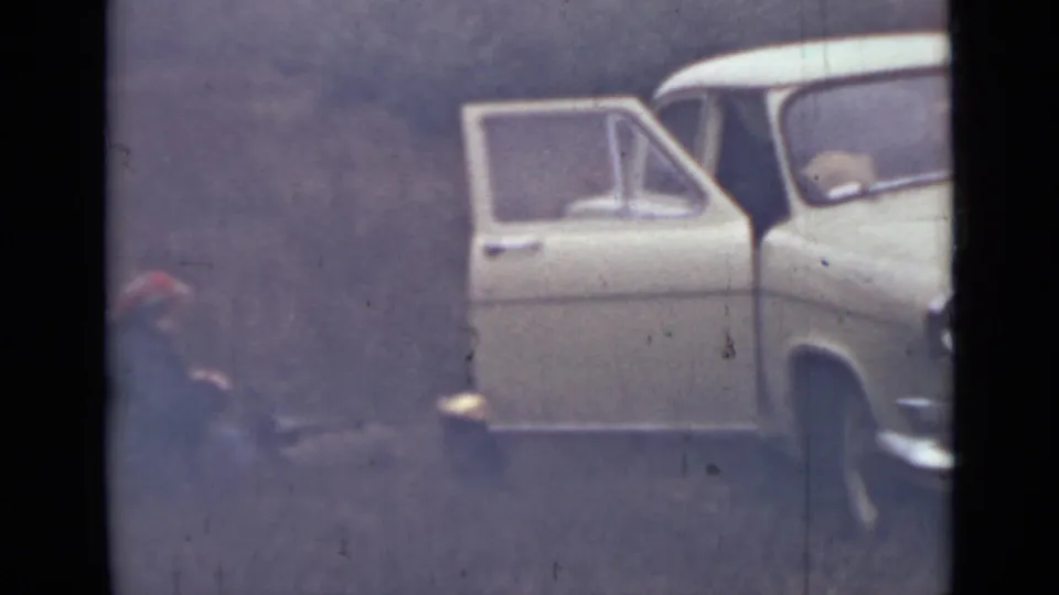 Soft 8mm frame from 1967 Saint Petersburg showing a crouching figure beside an open white car in a wooded or rural setting.