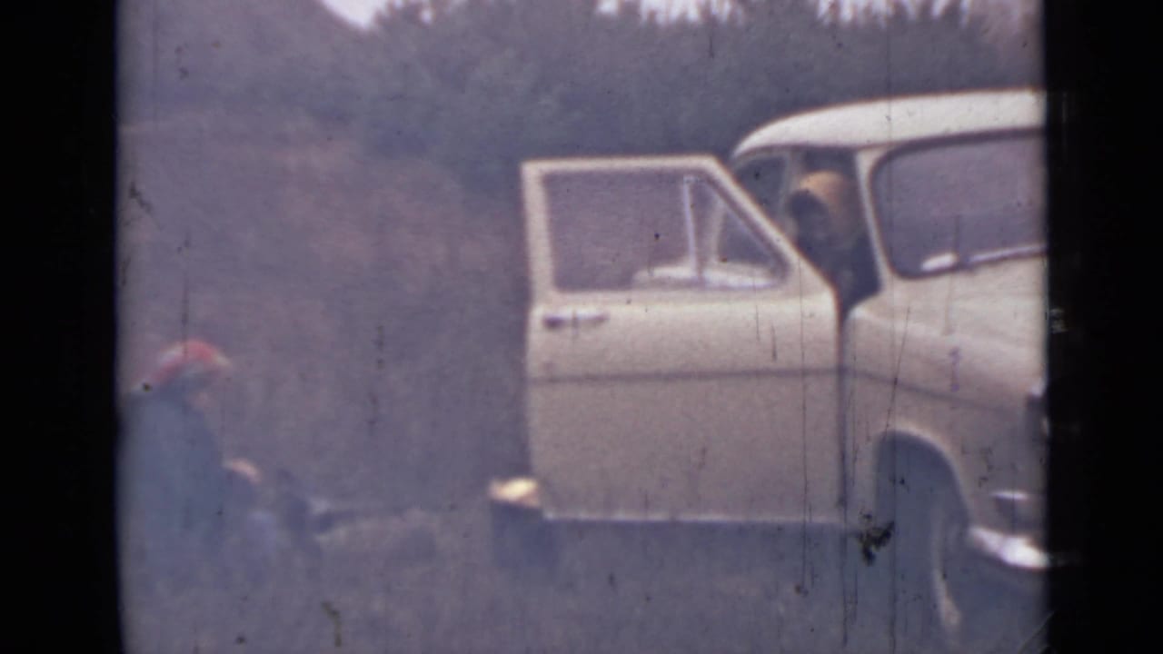 Light-colored car with open door parked in woods, with a seated person in a red hat nearby.