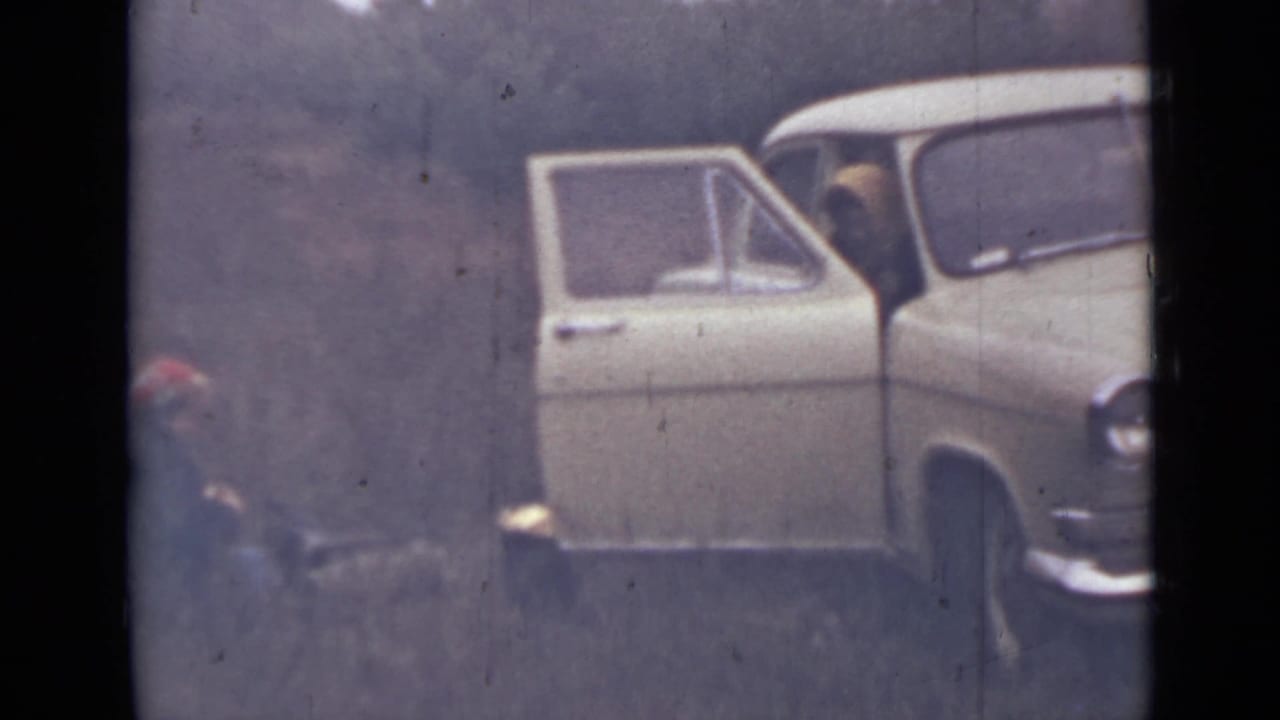 Open car door and front side of a pale car beside a wooded slope, with a person low at left.