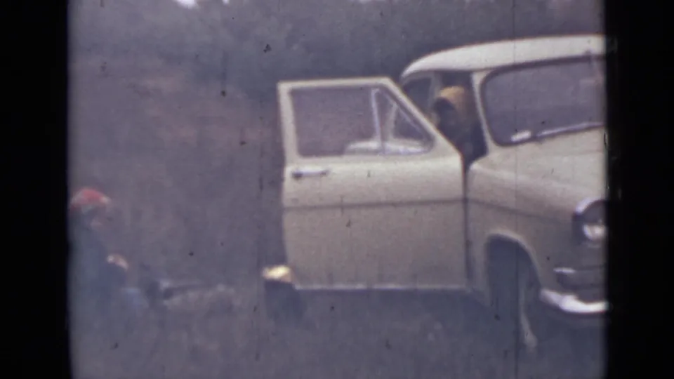 1967 home-movie frame near Saint Petersburg with an open car door, wooded hillside, and a person partly visible near the ground at left.