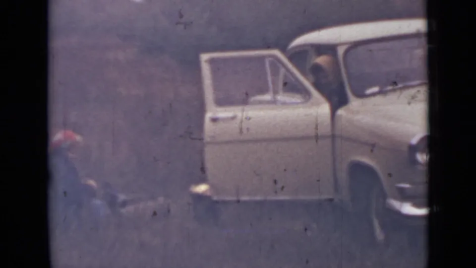 1967 8mm frame near Saint Petersburg showing the side of a pale car with its door open and a seated person in a red hat nearby.