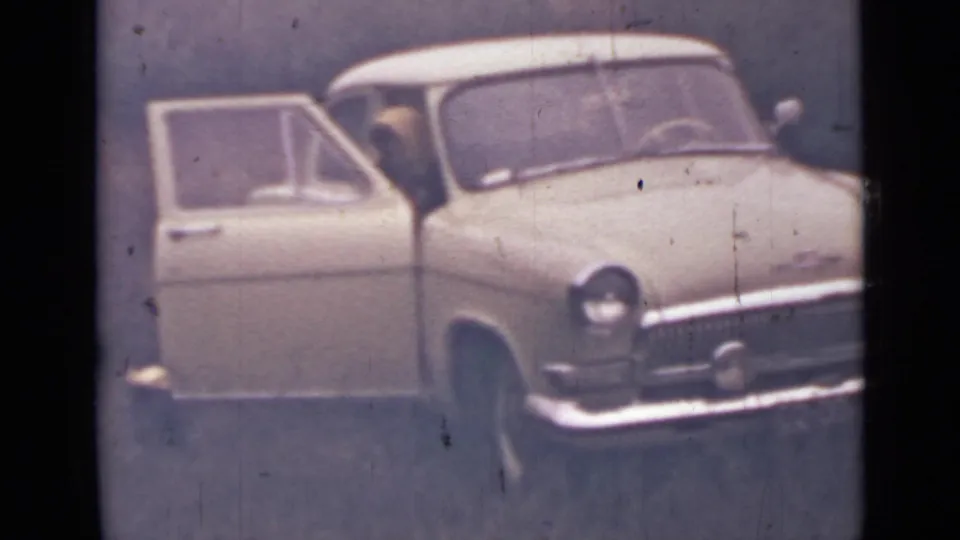 1967 home-movie frame near Saint Petersburg showing a pale classic car in the woods with its driver-side door open and a figure partly visible inside.