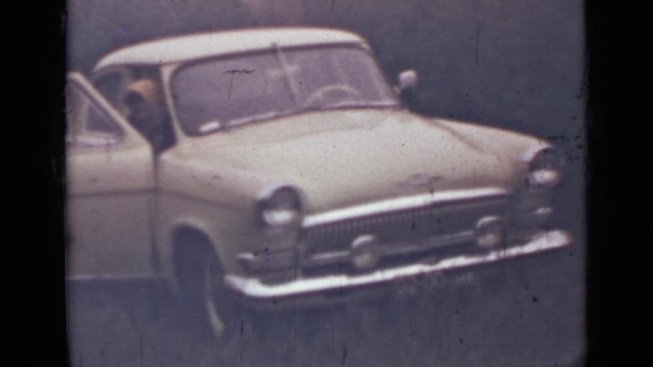 Older light-colored car in a wooded area with the driver door open.