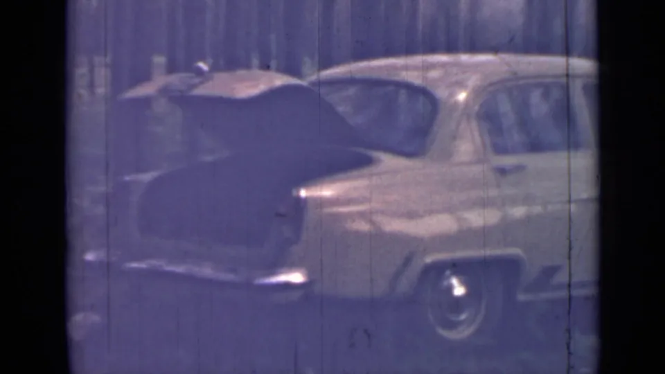 1967 8mm frame showing an older sedan in a wooded clearing, its trunk raised and side profile partly visible.