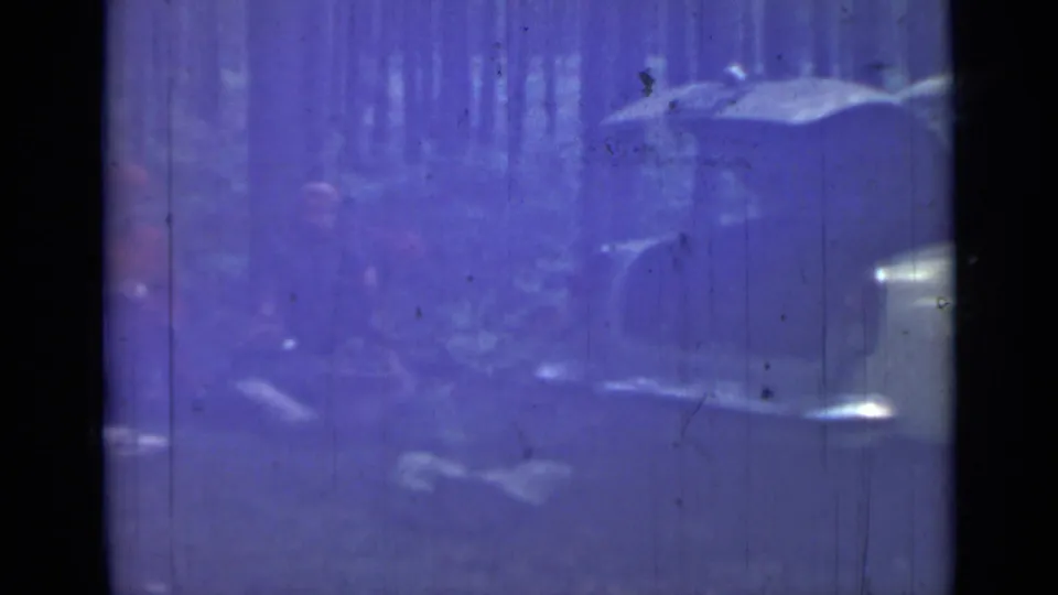 1967 8mm frame with a soft, purple-tinted view of people and belongings near a parked car in a forest setting.