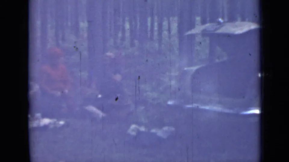 Aged 8mm frame from the forest outing, with seated figures in the background and part of an older pale sedan entering at right.