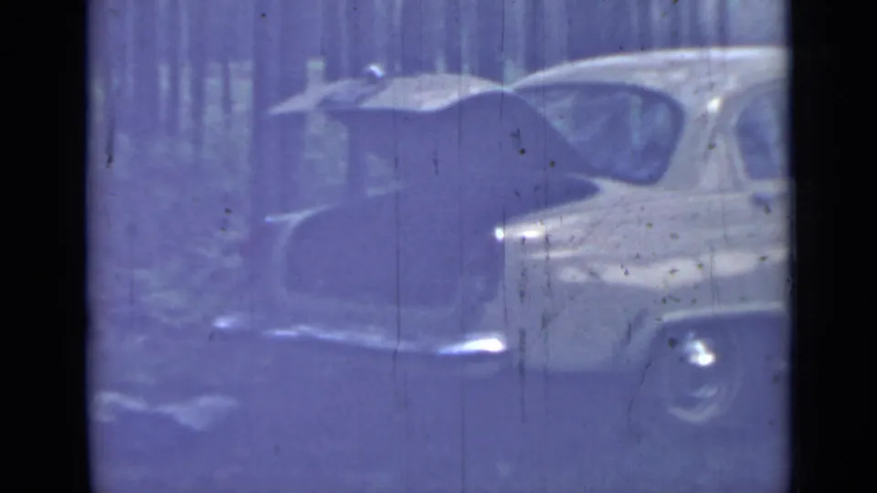 1967 8mm home-movie frame showing an older pale sedan parked in a wooded area near Saint Petersburg, with strong color fading and wear marks.