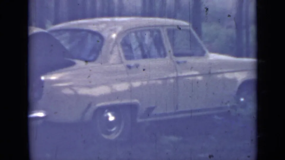 Side view of a pale older sedan in the wooded 1967 home-movie scene, framed by dark film edges and visible 8mm surface wear.