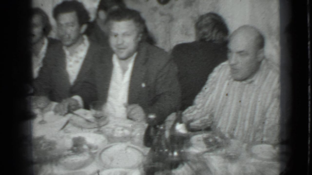 Several adult men sit around a dining table with plates, glasses, and bottles.