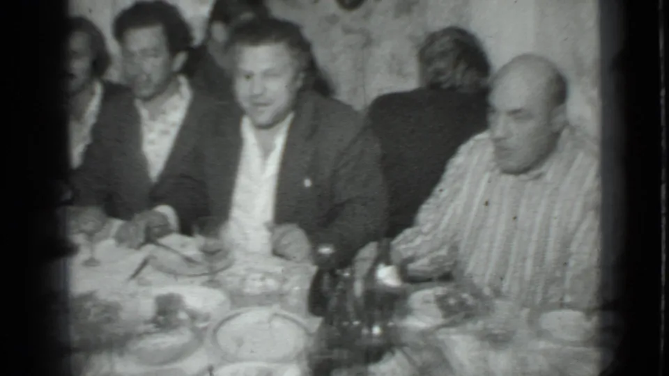 Black-and-white 8mm frame from a 1975 Moscow home movie showing adult men seated at a crowded dinner table.