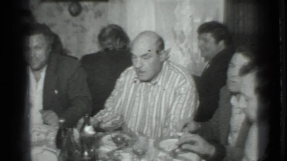 A 1975 Moscow home-movie frame shows men seated at dinner, with bottles and plates visible on the table.