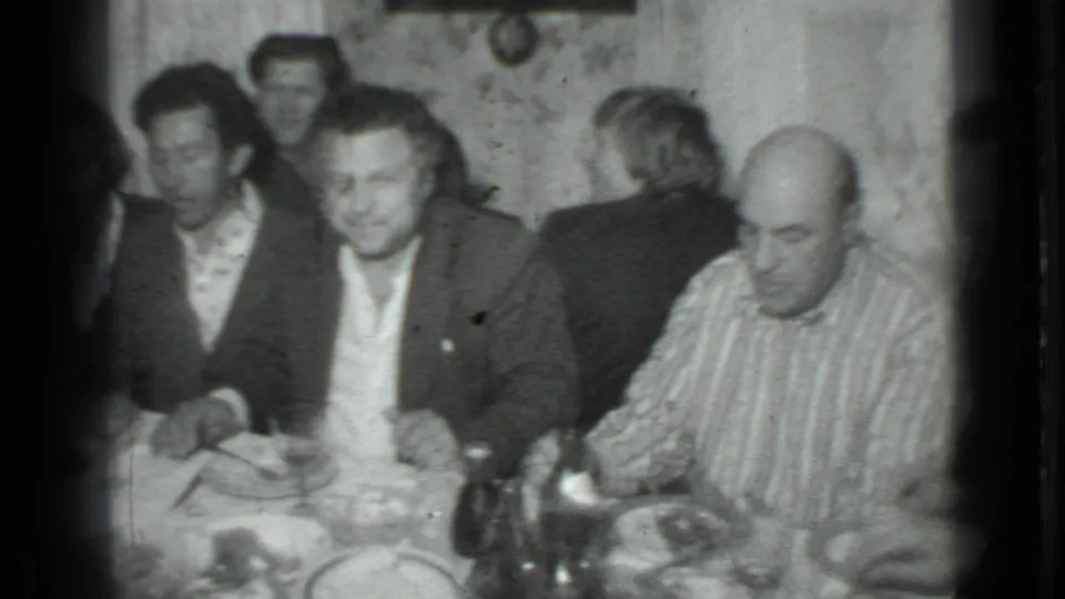 Black-and-white 8mm still of a private Moscow dinner scene with men seated near plates, glasses, and bottles.