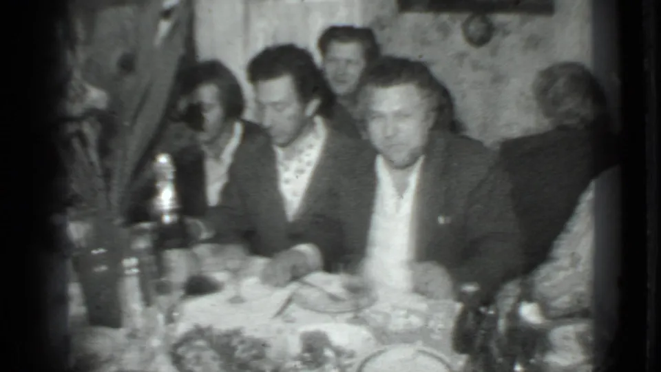 A 1975 Moscow home-movie frame shows a group of adult men at a private dining table in soft black-and-white 8mm footage.
