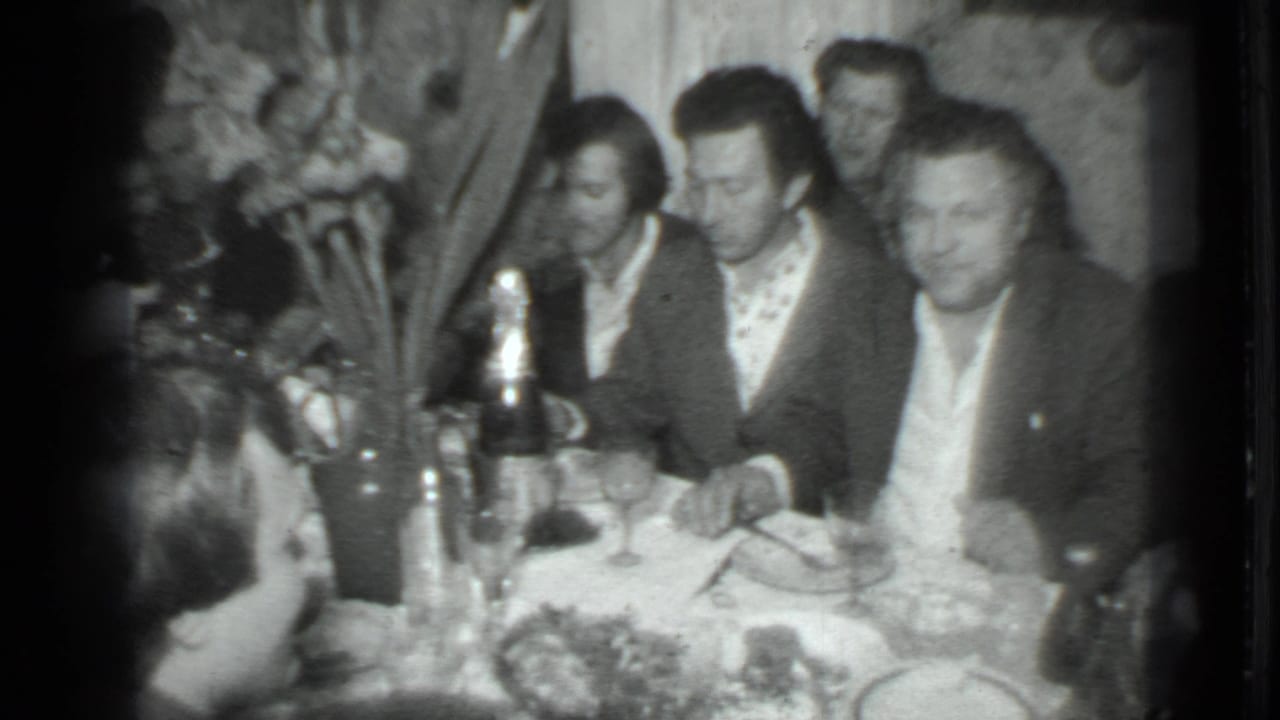 Men seated around a dinner table with bottles, glasses, food, and floral decor.
