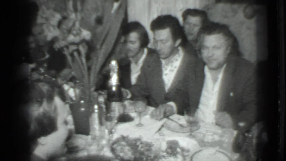 1975 Moscow home-movie frame showing men in suits and shirts seated at a crowded dinner table in a private interior.