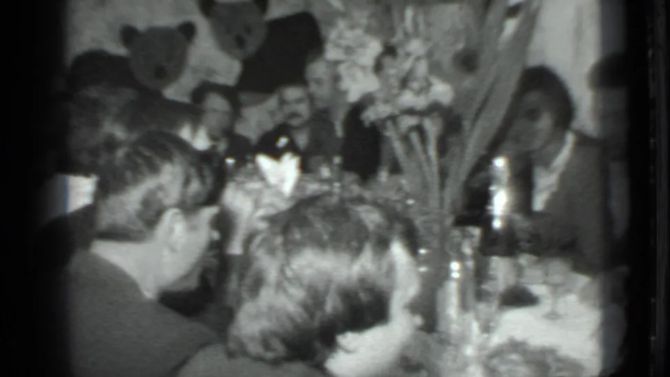 Handheld 1975 Moscow interior view across a crowded dinner table toward men seated beneath wall decorations.