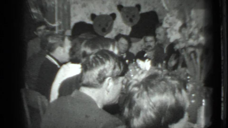 A 1975 Moscow home-movie frame shows guests from behind as men converse across a dining table in a private room.
