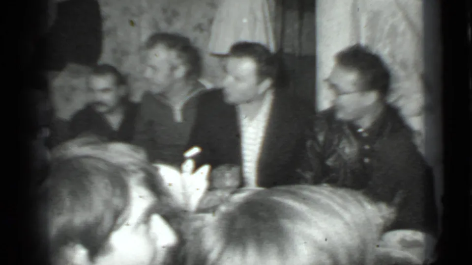 1975 Moscow home-movie frame of adults seated around a dinner table, with faces in profile and a handheld black-and-white film look.