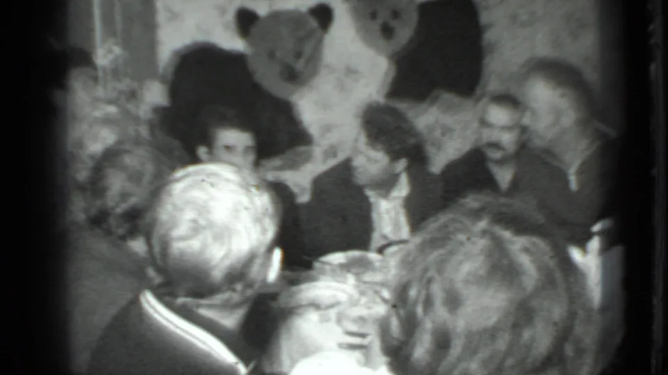 Adults sit and talk around a table in a Moscow home interior, with patterned wallpaper and a large bear wall decoration visible behind them.