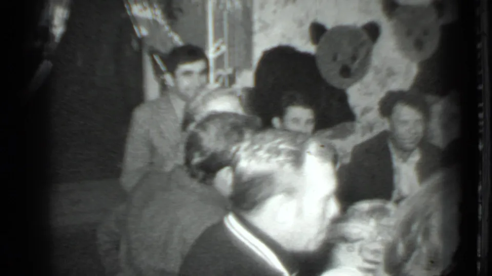 Black-and-white 8mm frame from a 1975 Moscow gathering, showing adults around a dinner table with a large bear decoration in the background.