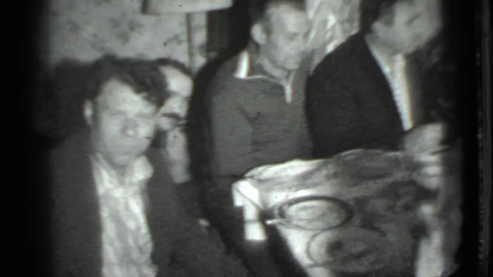 A close 1975 Moscow home-movie view shows adults seated beside a table, with dishes and a private apartment interior visible.