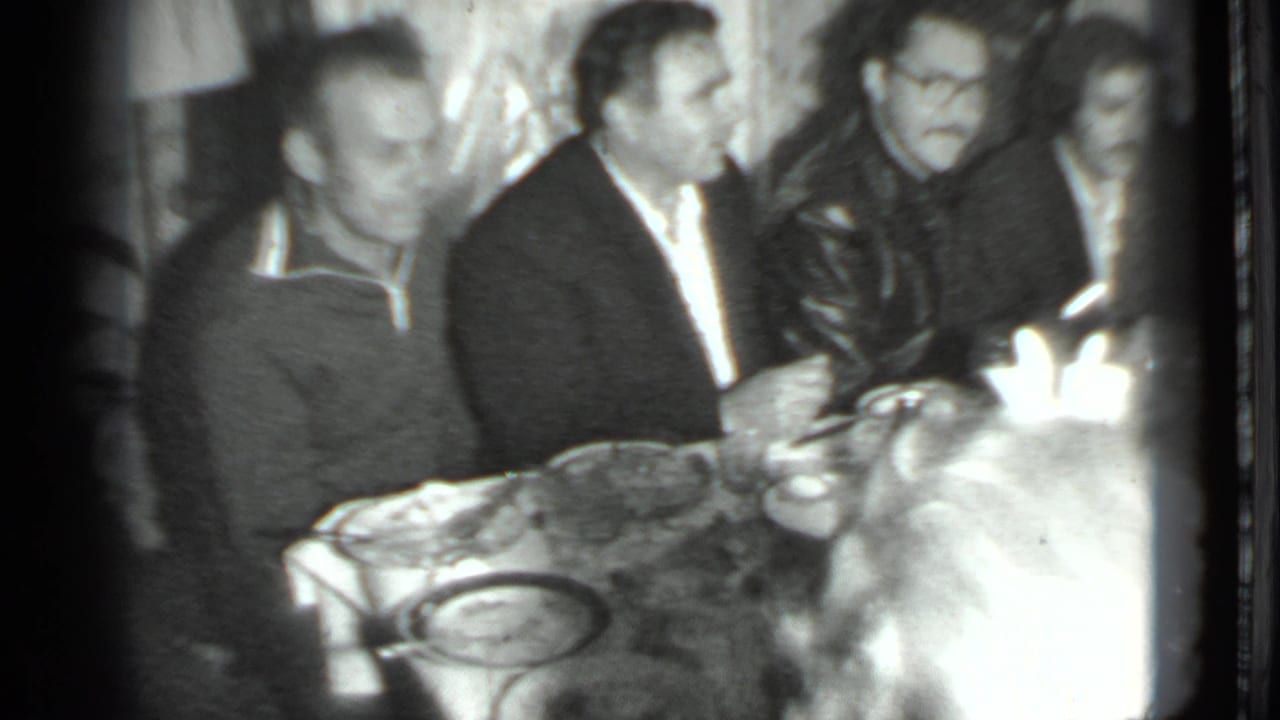 1975 Moscow 8mm frame showing several men gathered at a dinner table, with tableware and bottles visible in soft black-and-white footage.
