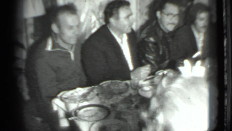 1975 Moscow 8mm frame showing several men gathered at a dinner table, with tableware and bottles visible in soft black-and-white footage.