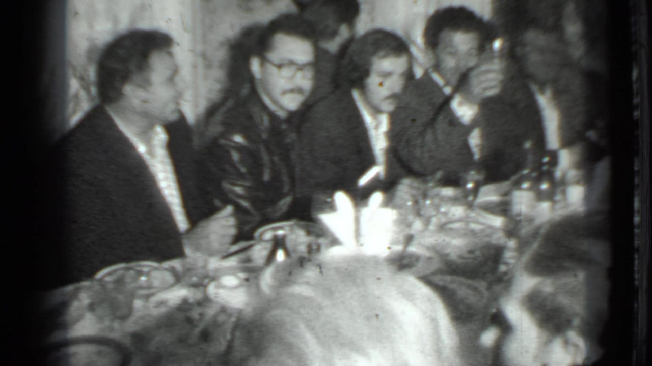 Group of men at a table, one man facing another while others look on.