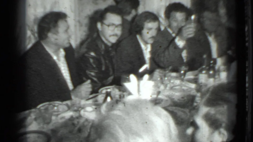 Black-and-white 8mm frame from the Moscow dinner scene, with men seated together around bottles, plates, and glasses.