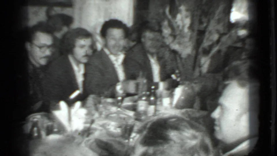 1975 Moscow home-movie frame showing men at a crowded dining table, with soft focus, grain, bottles, and plates visible.