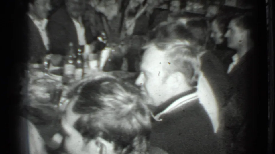 Black-and-white 8mm frame from the Moscow gathering, with foreground profiles and a table filled with bottles, glasses, and plates.