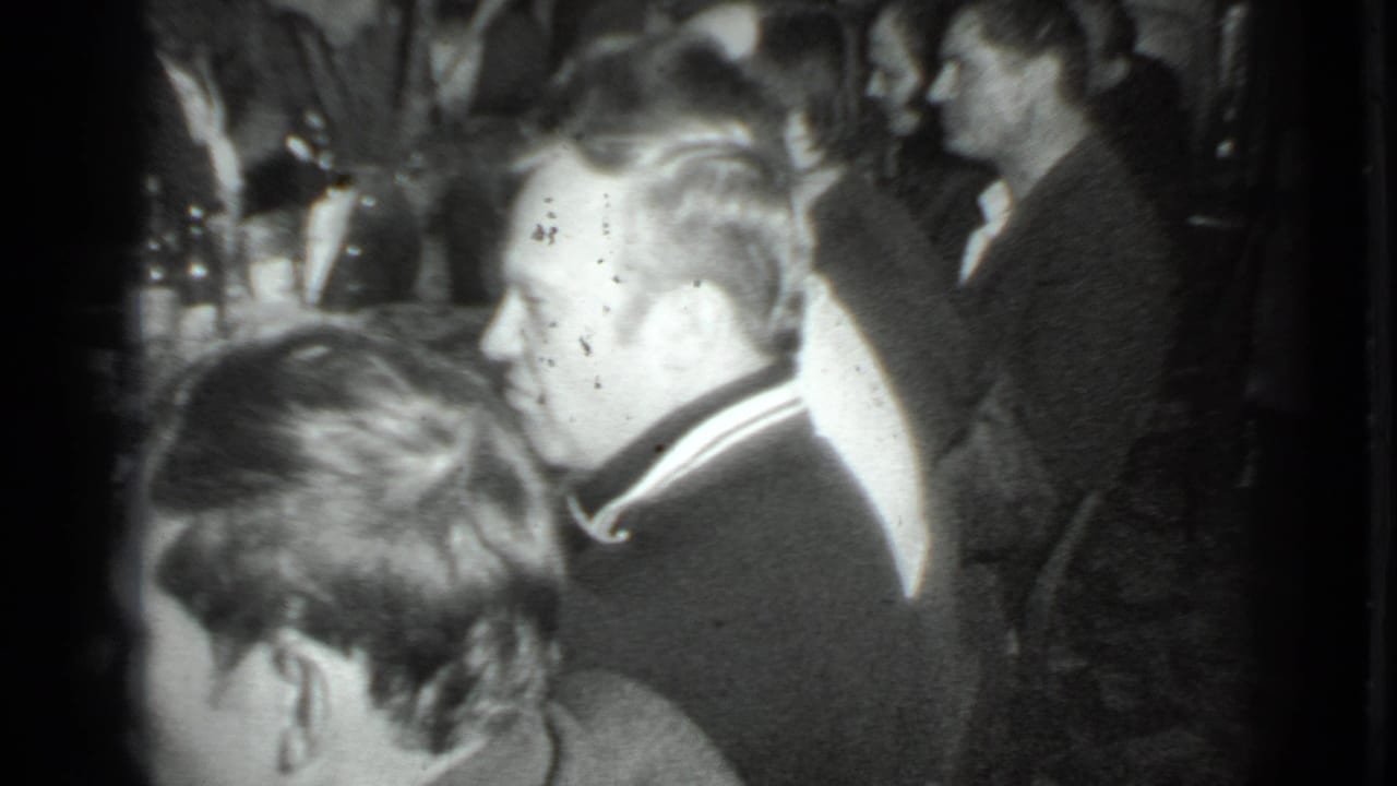 1975 Moscow home-movie frame showing adults in suits gathered in a dim private interior during a dinner conversation.