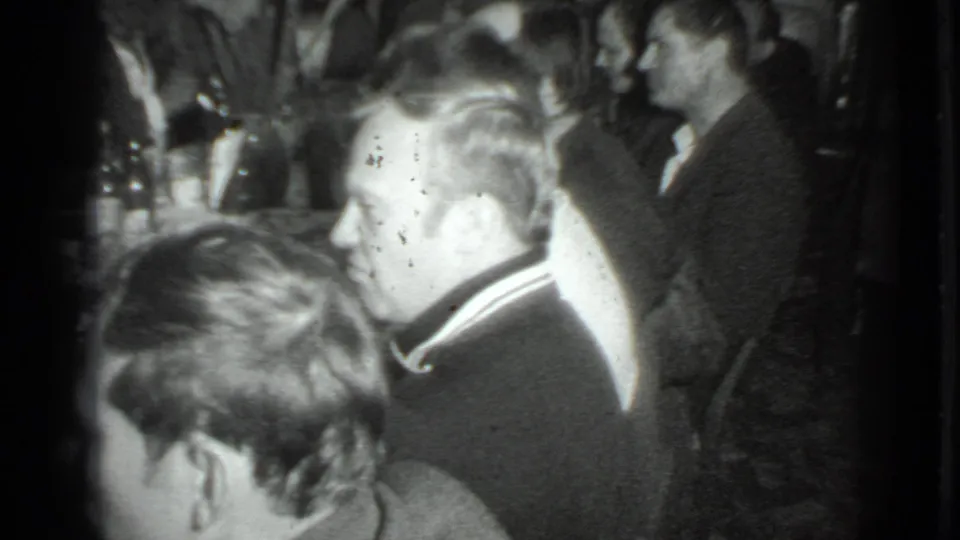 1975 Moscow home-movie frame showing adults in suits gathered in a dim private interior during a dinner conversation.