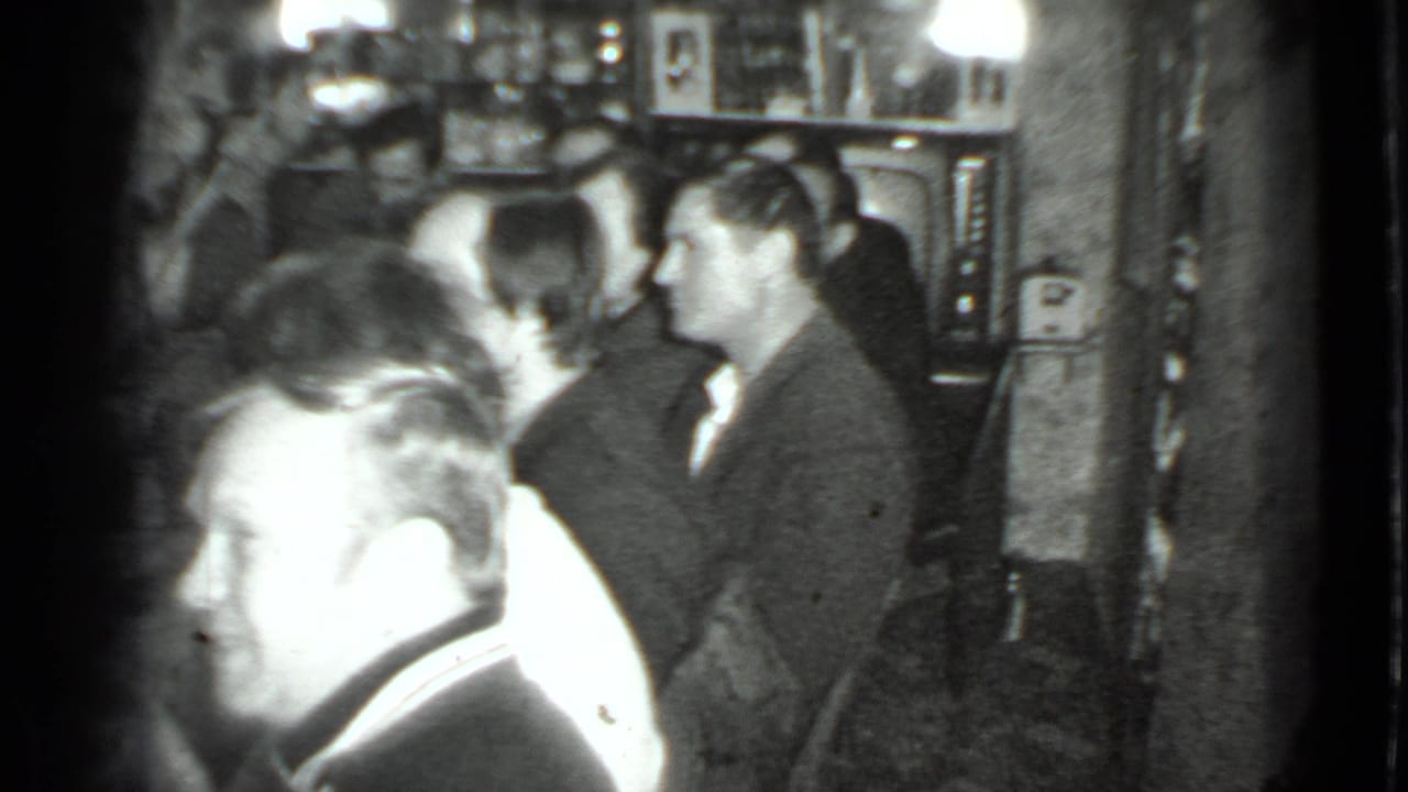 Home-movie frame from the Moscow dinner showing adults talking in a compact room with bottles, shelves, and a television nearby.