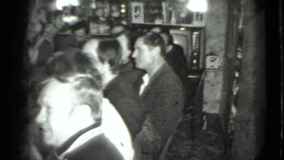 Home-movie frame from the Moscow dinner showing adults talking in a compact room with bottles, shelves, and a television nearby.