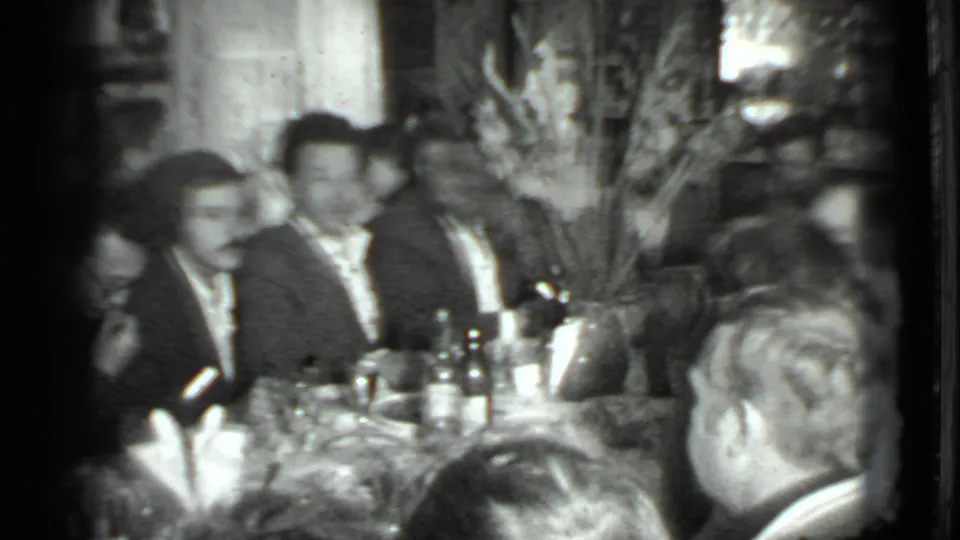 Black-and-white 8mm frame from the Moscow 1975 clip showing adults in suits seated at a dinner table during a private gathering.
