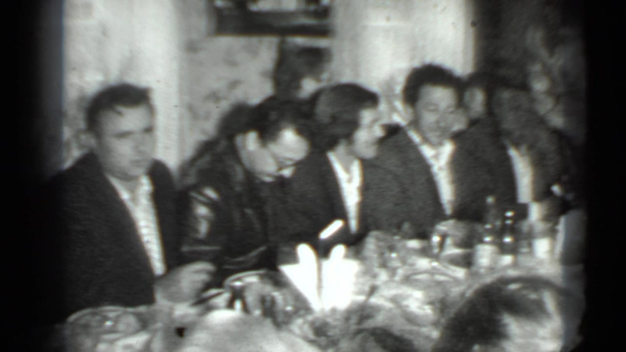 Dinner guests sit around a table with bottles, glasses, plates, and a bright table decoration.