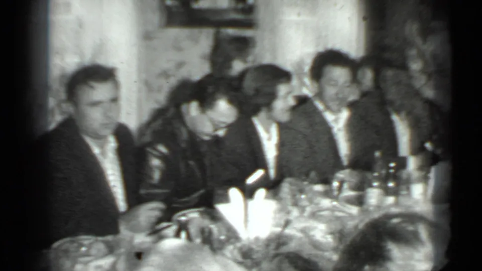 Moscow 1975 home-movie frame showing adults gathered at a dinner table, some turned toward conversation in a private room.