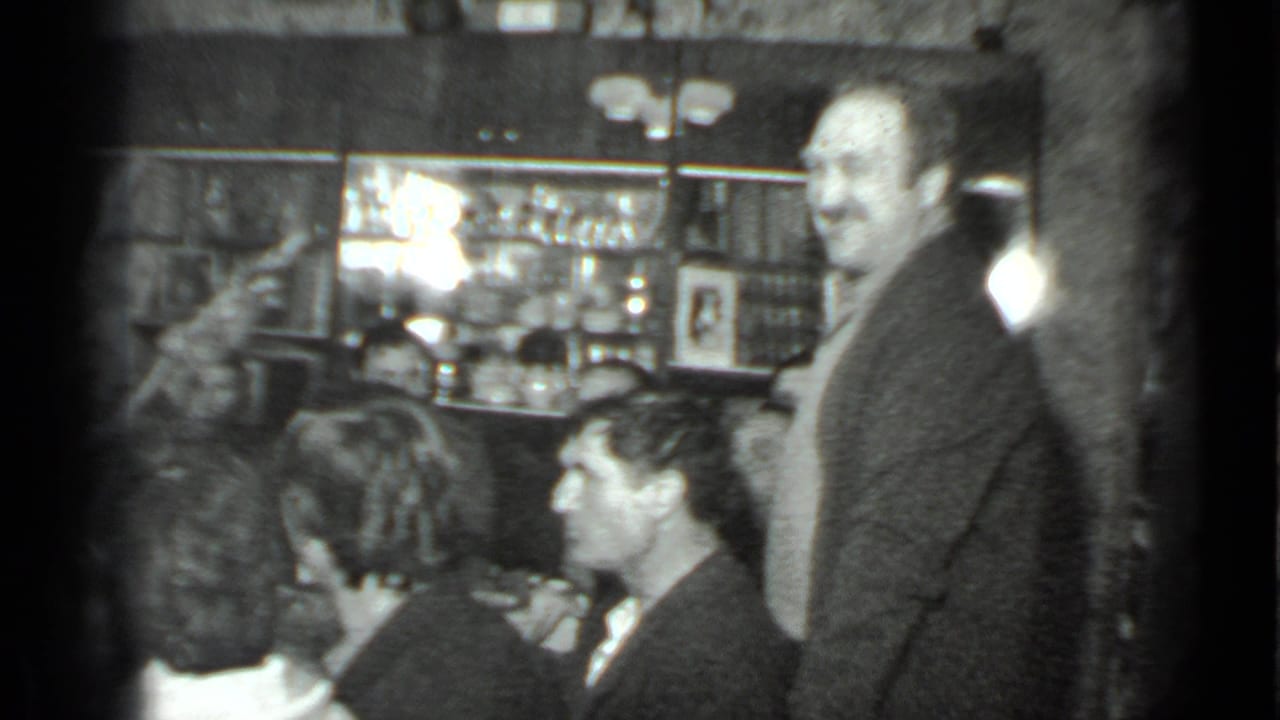 Black-and-white 8mm frame from the 1975 Moscow gathering showing a man standing near seated guests with bar shelves in the background.