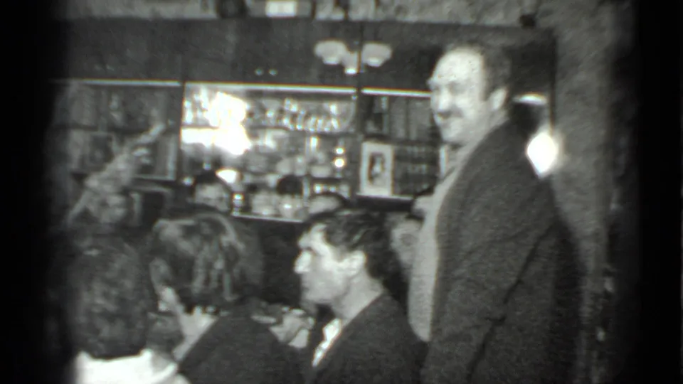 Black-and-white 8mm frame from the 1975 Moscow gathering showing a man standing near seated guests with bar shelves in the background.