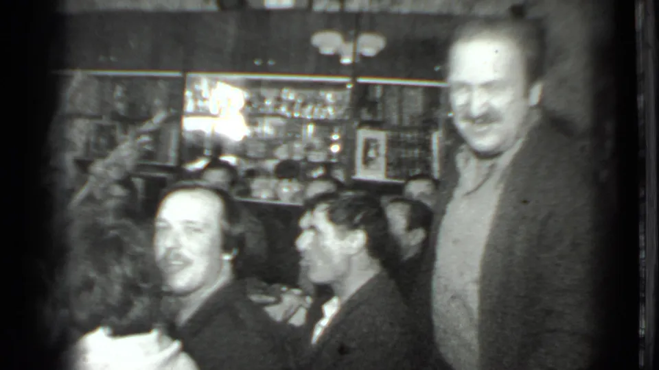 Moscow 1975 home-movie frame showing a smiling man standing by a seated group during an indoor dinner gathering.