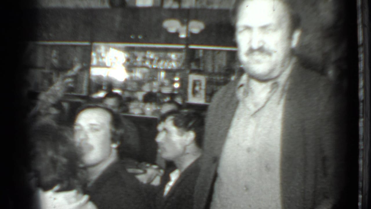 Crowded interior with seated men, a standing man, shelves, bottles, and glassware.