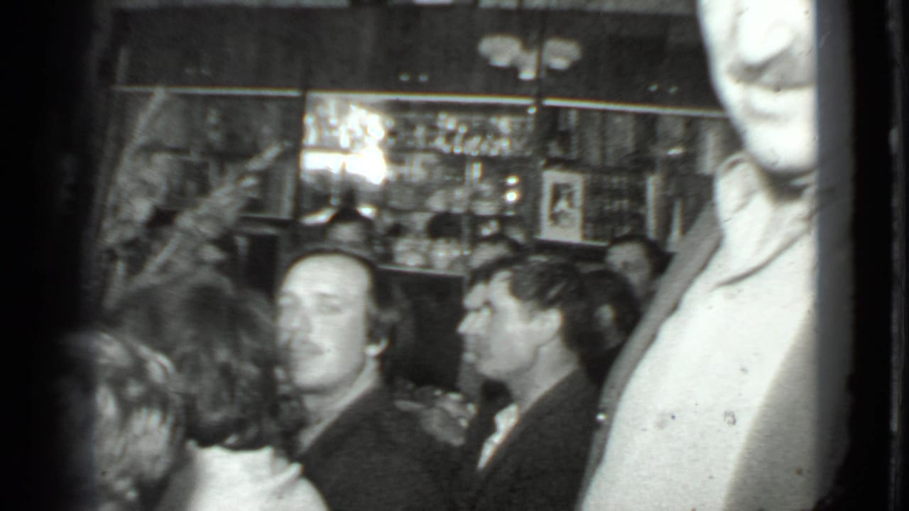 Adults sit closely together while a woman stands near shelves and glassware.
