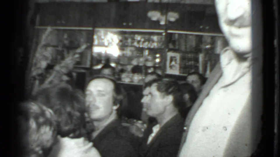 1975 Moscow home-movie frame with seated adults in conversation and a standing woman beside shelves of glassware.