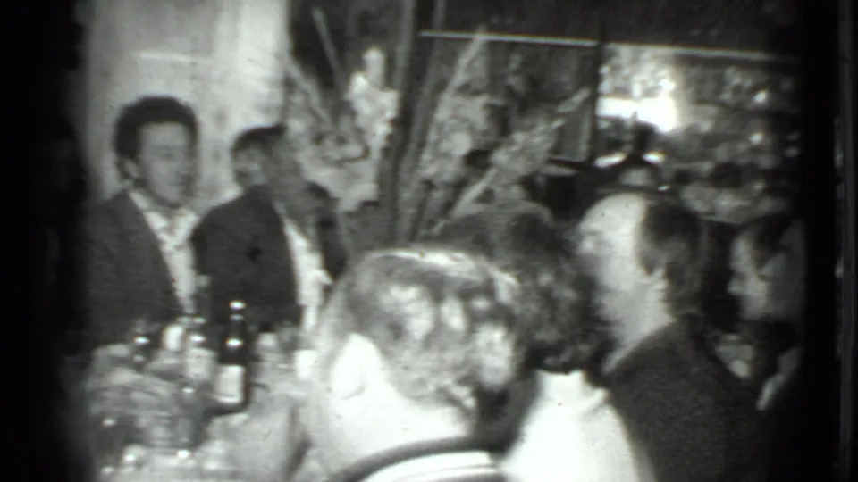 1975 Moscow home-movie frame showing a long table cluttered with bottles and glasses as adults gather around it.