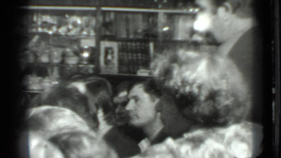 1975 Moscow home-movie frame with tightly framed adults gathered in conversation near shelves and wall decor.