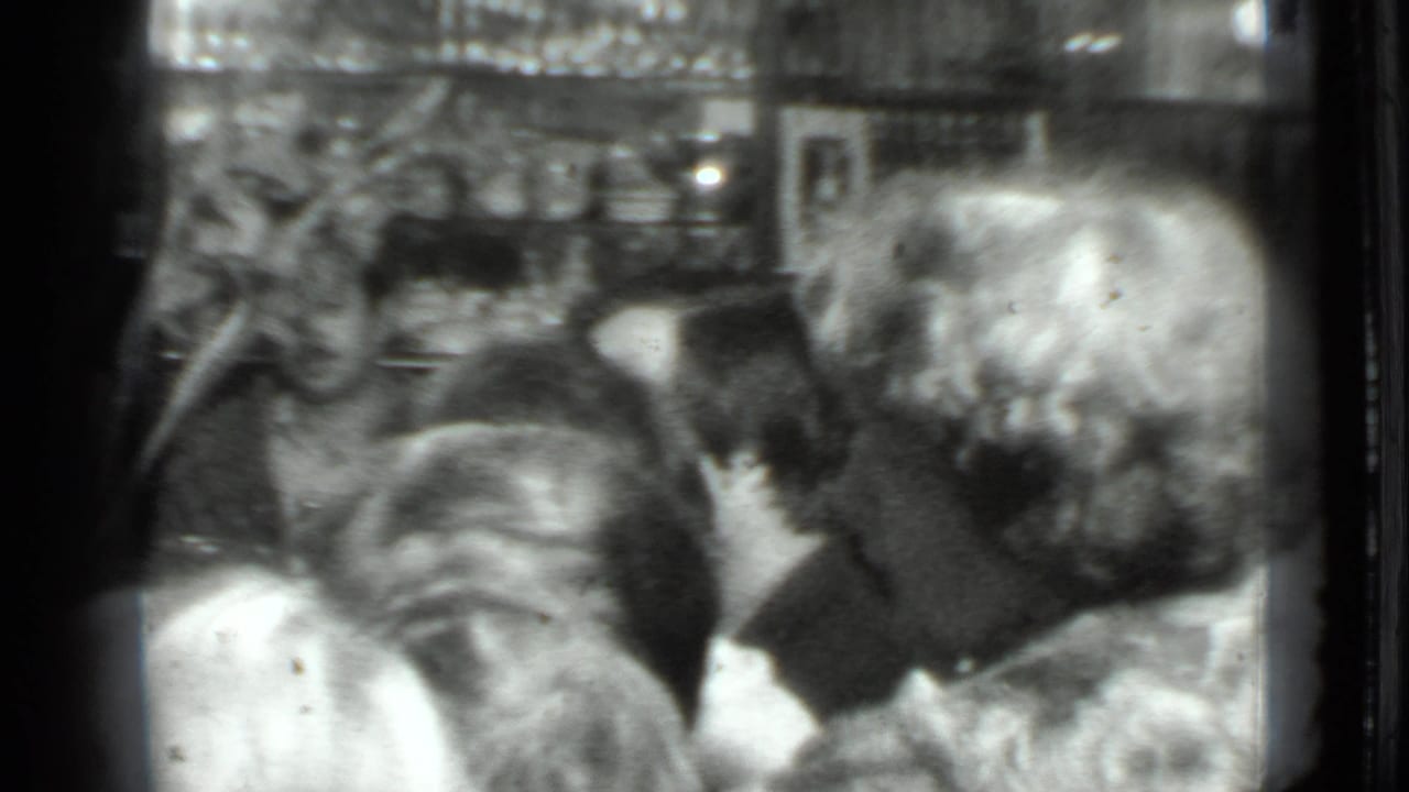 Grainy 8mm frame of adults at a crowded Moscow dinner table, with heads turned toward conversation and food partly visible.